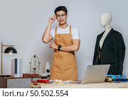 Cheerful young Asian fashion designer or tailor wearing eyeglasses use laptop computer to search idea or online orders, mannequin in suit and full of tailoring tools with equipment on desk in studio. Стоковое фото, агентство Ingram Publishing / Фотобанк Лори
