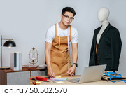 Cheerful young Asian fashion designer or tailor wearing eyeglasses use laptop computer to search idea or online orders, mannequin in suit and full of tailoring tools with equipment on desk in studio. Стоковое фото, агентство Ingram Publishing / Фотобанк Лори
