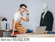 Cheerful young Asian fashion designer or tailor wearing eyeglasses use laptop computer to search idea or online orders, mannequin in suit and full of tailoring tools with equipment on desk in studio. Стоковое фото, агентство Ingram Publishing / Фотобанк Лори