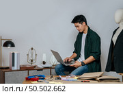 Portrait Young handsome male designer use laptop computer for searching good idea to design fashion clothing, mannequin and equipment with full of tailoring tools on desk in studio. Стоковое фото, агентство Ingram Publishing / Фотобанк Лори