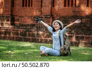 Cheerful young backpacker sitting on grass raise up her arm and smiling with happiness, holding a camera in hand while traveling in ancient site. Стоковое фото, агентство Ingram Publishing / Фотобанк Лори