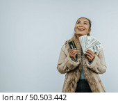Portrait of successful Asian woman with a fur coat holding money and card, smiling and looking aside, standing over background in studio with copy space. Advertising concept. Стоковое фото, агентство Ingram Publishing / Фотобанк Лори