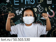 Portrait of a little girl wearing eyeglasses and facemask at school to avoid the spread of coronavirus, new normal with schoolchild,  blure blackboard in background. Стоковое фото, агентство Ingram Publishing / Фотобанк Лори