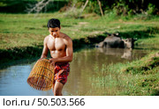 Portrait Young man topless use bamboo fishing trap to catch fish for cooking, Asian young farmer man in rural lifestyle, blurred buffalo is soaking in the swamp. in background. Стоковое фото, агентство Ingram Publishing / Фотобанк Лори
