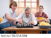 In the living room of the house, an elderly couple sits and relaxes. to begin playing chess together with a chess board with a daughter cheering beside him. Стоковое фото, агентство Ingram Publishing / Фотобанк Лори