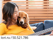 On a weekend, A young Asian woman and her beagle puppy are playing the guitar and singing cheerfully together in the living room of a house. Стоковое фото, агентство Ingram Publishing / Фотобанк Лори
