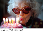 An elderly woman wearing sunglasses blows out candles on a birthday cake, smiling and celebrating her special day. Стоковое AI-изображение, AI-художник Катерина Бондарец / Фотобанк Лори