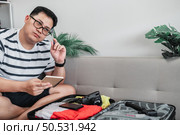 Asian man glasses is preparing clothes in suitcases. He’s choosing clothes, travel documents, the itinerary for a solo trip, and check the checklist. Travel, holiday and vacation concept. Стоковое фото, агентство Ingram Publishing / Фотобанк Лори