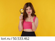 Portrait of detective pretty woman holding magnifying glass and pointing finger at camera isolated over yellow background. Investigation, discovery and vision concept. Стоковое фото, агентство Ingram Publishing / Фотобанк Лори