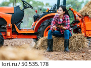 Asian young female farmer sitting on bale of hay with big tractor machine in farmland. Modern technologies in agriculture management and agribusiness concept. Стоковое фото, агентство Ingram Publishing / Фотобанк Лори