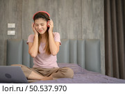 Asian woman puts on headphones and enjoys listening to music from laptop on the bed in the bedroom. Стоковое фото, агентство Ingram Publishing / Фотобанк Лори