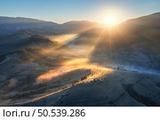 Aerial view of mountains and hills in low clouds at sunrise. Autumn in Ukraine. Top view of Carpathians mountains in fog in fall at dawn. Beautiful landscape with yellow meadows, forest, village, sun. Стоковое фото, агентство Ingram Publishing / Фотобанк Лори