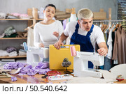 Focused young repairman repairing sewing-machine and Asian tailoress talking. Стоковое фото, фотограф Яков Филимонов / Фотобанк Лори