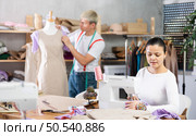 Young Asian tailoress using sewing-machine while other works with mannequin. Стоковое фото, фотограф Яков Филимонов / Фотобанк Лори