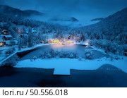 Aerial view of illuminated houses in fairy village in snow, forest, Jasna lake, street lights at winter night. Top view of alpine mountains in fog, snowy pine trees at dusk. Kranjska Gora, Slovenia. Стоковое фото, агентство Ingram Publishing / Фотобанк Лори