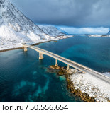 Aerial view of bridge over the sea and snowy mountains in Lofoten Islands, Norway. Top drone view of bridge in cold winter day. Wintry landscape with blue water, rocks in snow, road, cloudy sky. . Стоковое фото, агентство Ingram Publishing / Фотобанк Лори