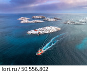 Aerial view of floating fishing boat in blue sea in snowy Henningsvaer village, Lofoten islands, Norway in winter. Top drone view of boat, mountains in snow, sea, cloudy sky, town, rorbuer and houses. Стоковое фото, агентство Ingram Publishing / Фотобанк Лори