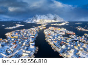 Aerial view of snowy Henningsvaer fishing village, Lofoten islands, Norway in winter at sunset. Top drone view of mountains in snow, sea, boats, bridge, road, cloudy sky, town, rorbuer and houses. Стоковое фото, агентство Ingram Publishing / Фотобанк Лори