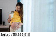 Pregnant asian woman holding glass of milk and smiles happily talking to her unborn child. Стоковое фото, агентство Ingram Publishing / Фотобанк Лори