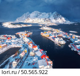 Aerial view of snowy Henningsvaer fishing village, Lofoten islands, Norway in winter at sunset. Top drone view of mountains in snow, sea, boats, bridge, road, cloudy sky, town, rorbuer and houses. Aerial view of snowy fishing village in Norway in winter. Стоковое фото, агентство Ingram Publishing / Фотобанк Лори