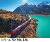 Aerial view of modern red train near azure mountain Bianco lake, snowy rocks and blue sky at sunset in autumn. Bernina Express, Switzerland. Top view of train in Alps, railroad, orange hills in fall. Стоковое фото, агентство Ingram Publishing / Фотобанк Лори