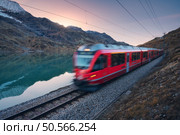 Moving modern red train near azure mountain Bianco lake, snowy rocks and colorful sky at sunset in autumn. Bernina Express, Switzerland. Blurred train in Alps, reflection in water in fall at dusk. Стоковое фото, агентство Ingram Publishing / Фотобанк Лори