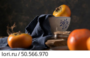Ripe persimmons on a navy cloth with rustic wooden elements create a cozy autumn scene. Soft lighting highlights the fruit’s vibrant color and texture. Стоковое фото, агентство Ingram Publishing / Фотобанк Лори