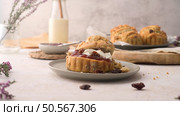 Rustic scone with cream and jam on a plate, surrounded by raisins, flowers, and a milk bottle in a cozy, soft-lit setting. Стоковое фото, агентство Ingram Publishing / Фотобанк Лори