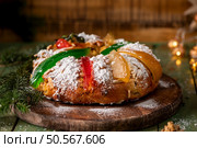 Festive rosca de reyes bread topped with candied fruit and powdered sugar on wooden board with pine branches and twinkling lights in background. Стоковое AI-изображение, агентство Ingram Publishing / Фотобанк Лори
