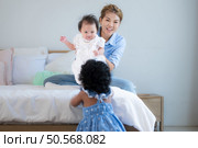A small girl stood at the foot of the bed playing with the younger sibling carried by his mother and smiling with affection, In a bedroom surrounded by warmth and family. Стоковое фото, агентство Ingram Publishing / Фотобанк Лори