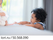 A small girl stood at the foot of the bed playing with the younger sibling carried by his mother and smiling with affection, In a bedroom surrounded by warmth and family. Стоковое фото, агентство Ingram Publishing / Фотобанк Лори