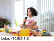 A tiny girl sits on a toy table, clutching a microphone, and sings cheerfully in the living room. Стоковое фото, агентство Ingram Publishing / Фотобанк Лори