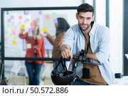 Portrait of smiling businessman riding electric scooter in office with colleague in background. Стоковое фото, агентство Ingram Publishing / Фотобанк Лори