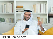 Portrait of a smiling Arabian man holding a glass of champagne and using mobile phone at home. Стоковое фото, агентство Ingram Publishing / Фотобанк Лори