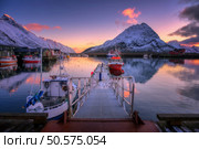 Red fishing boats on the sea, reflection in water, snowy mountains, colorful purple sky with pink clouds at sunset. Lofoten islands, Norway. Winter landscape with boats in harbor, fjord, rorbu at dusk. Стоковое фото, агентство Ingram Publishing / Фотобанк Лори
