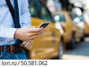 Close up male hand calling a taxi with a smartphone app. Close up of a businessman using mobile phone. Closeup detail of a businessman’s hand holding a cellphone with yellow taxi in background. . Стоковое фото, агентство Ingram Publishing / Фотобанк Лори