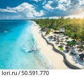 Aerial view of the sailboat on blue sea, empty white sandy beach, umbrellas at sunset. Summer in Zanzibar. Tropical scenery with boat, ocean with waves, green palms, sky. Top drone view. Exotic. Стоковое фото, агентство Ingram Publishing / Фотобанк Лори