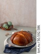 A freshly baked Portuguese folar cake sits on a plate with a nest containing speckled green Easter eggs and delicate feathers scattered around. Стоковое фото, агентство Ingram Publishing / Фотобанк Лори