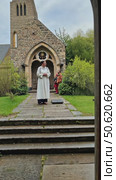 A female priest in a white cassock stands outside a historic stone church, viewed through an arched doorway. Стоковое AI-видео, аниматор Sergey Grishin / Фотобанк Лори