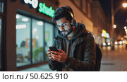 Hispanic young attractive man stands in dark street in front of shop, changes songs and tracks on smartphone, listens to music in wireless headphones. Hipster wearing casual dress. Стоковое AI-видео, аниматор Sergey Grishin / Фотобанк Лори