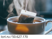 Teabag above a white ceramic mug with a steaming drink. Hand is picking a tea bag into a cup of tea. Teabag in hot water. Morning beverage. Tagged teabags with string. Someone preparing tea. Стоковое AI-изображение, AI-художник Марина Демидюк / Фотобанк Лори