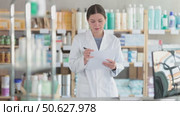 Skilled young female pharmacy employee in white coat standing with checklist, verifying product availability on shelves during routine inventory control. Стоковое видео, видеограф Яков Филимонов / Фотобанк Лори