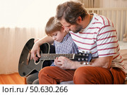 Handsome dad teaches his cute son to play the guitar sitting on sofa in the living room at home. Happy Father's Day. Father teaching his child to play guitar. Kid Learning music skill. Art education. Стоковое фото, фотограф Марина Демидюк / Фотобанк Лори