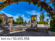 The image shows one of the central sites of the New Chersonesus cultural and historical complex in Sevastopol. The frame shows two monumental icons depicting Jesus Christ and the Virgin Mary with the baby, placed in decorative arches decorated with fresh flowers. The space is paved with ornamental stone tiles, in the background are neat rows of trees, decorative lamps and a Byzantine-style building. The blue sky with cirrus clouds gives the composition a solemn and calm feel. Стоковое фото, фотограф Анна Кривицкая / Фотобанк Лори