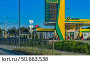Summer day in the south of Russia, Crimea. The picture shows a modern gas station "ATAN" with a bright corporate design in green and yellow tones. The electronic board shows the current prices for gasoline, diesel and gas. In the frame - a clear blue sky, a well-kept roadside area with green spaces, a metal fence and road signs. The atmosphere of calm and order emphasizes the developed infrastructure and convenience of car travel in Crimea. In the distance, trees and elements of urban development are visible. Редакционное фото, фотограф Анна Кривицкая / Фотобанк Лори