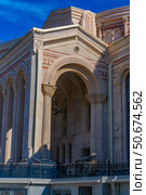 An architectural fragment of the building of the cultural and historical complex "New Chersonesos" in Sevastopol. In the foreground is a massive arch with decorative carvings and columns made of light stone. The architecture contains elements of the Byzantine and neoclassical styles: arched windows, geometric ornamentation, panels and carved capitals. Clear sunlight emphasizes the relief of the details. In the background is a bright blue sky, creating a visual contrast with the beige and pink palette of the facade. The building looks monumental, emphasizing the connection with the cultural heritage of Chersonesos. Стоковое фото, фотограф Анна Кривицкая / Фотобанк Лори