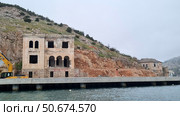 The photo shows the coast of Balaklava in Crimea with views of old ruined buildings built into a rocky slope. The water quietly splashes near the concrete embankment, on which construction equipment is visible, indicating restoration or construction work. The architecture of the dilapidated building with arches and massive masonry speaks of its historical past. Above all this hang cliffs overgrown with low vegetation. The atmosphere is slightly foggy, creating a feeling of abandonment, silence and time stopped by the sea. Стоковое фото, фотограф Анна Кривицкая / Фотобанк Лори