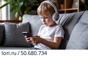 Cute blonde boy is sitting on the couch and looking at the phone. Стоковое AI-изображение, AI-художник Ирина Гильмуллина / Фотобанк Лори