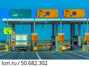Toll booth on the M4 Don toll road in Russia. In the foreground, a truck passes through a barrier in a special lane with a green arrow, intended for transponders (automated payment system). Above each lane, there are electronic boards with payment methods (cash, card, transponder) and accessibility information. The infrastructure is clearly organized: road signs, fences, CCTV cameras, and safety signs. In the background, there is an open rural landscape with fields and trees on the horizon. (2020 год). Редакционное фото, фотограф Анна Кривицкая / Фотобанк Лори