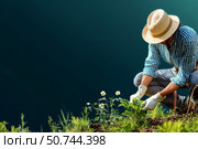 Gardener Tending to Flowers by a Tranquil Body of Water in a Sunny Outdoor Setting. Стоковое AI-изображение, AI-художник Юлия Гапеенко / Фотобанк Лори